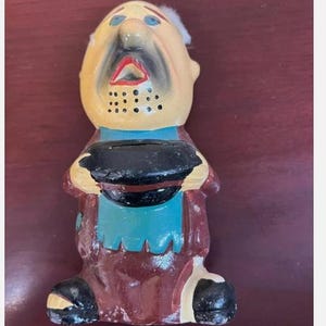 May include: A vintage ceramic piggy bank shaped like a man with a white beard, blue eyes, and a black mustache. He is wearing a brown coat, a blue shirt, and a black hat. The piggy bank has a slot on the back for coins.