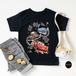 a black t-shirt with a graphic of three cartoon cars, a pair of gray jeans, a camera, and some other accessories, all laid out on a white background.