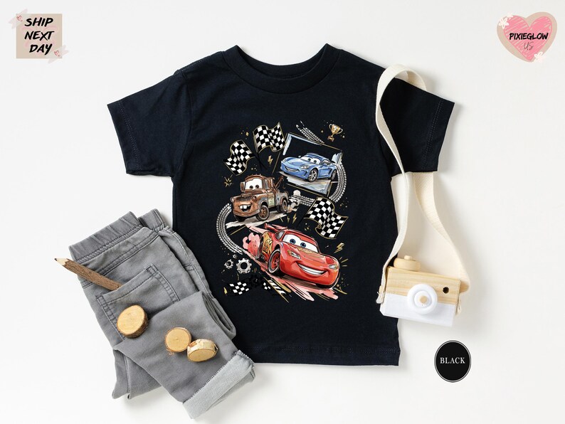 a black t-shirt with a graphic of three cartoon cars, a pair of gray jeans, a camera, and some other accessories, all laid out on a white background.