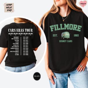 a woman wearing a black t-shirt with a Disney Cars-themed design. The t-shirt has a list of Disney Cars characters and a vintage-style van graphic. The woman is posing with the t-shirt, which appears to be a promotional or merchandise item for the Cars Eras Tour.