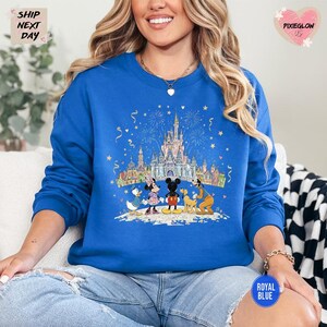 A woman wearing a blue sweatshirt with a Disney-themed graphic of Mickey Mouse, Minnie Mouse, Donald Duck, and Goofy on a castle, along with fireworks in the background.