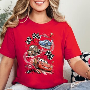 A woman wearing a red t-shirt with a graphic of three animated cars racing, including a blue car, a red car, and a brown truck, along with checkered flags and other racing elements.