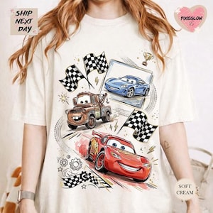 A white t-shirt with a graphic design featuring animated cars from the Disney Pixar movie Cars, including a red car, a blue car, and a brown truck. The design also includes checkered flags and other racing-themed elements.