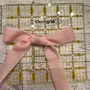 May include: A pink satin ribbon tied into a bow on top of a clear plastic ruler with yellow markings. The ruler is marked with inches and has a grid pattern. The text "Omnigrid" is printed on the ruler.