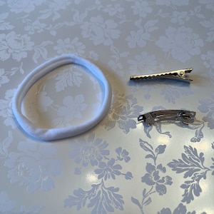 May include: A white fabric headband and two silver hair clips on a white floral background.