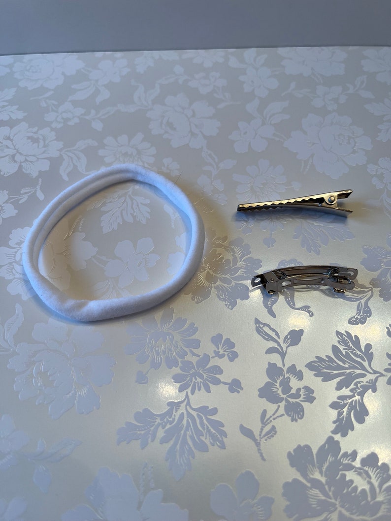 May include: A white fabric headband and two silver hair clips on a white floral background.