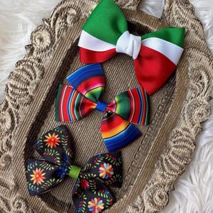 May include: Three decorative hair bows in a decorative tray. One bow features green, white, and red stripes. Another bow has a colorful striped pattern. The third bow is black with floral designs.