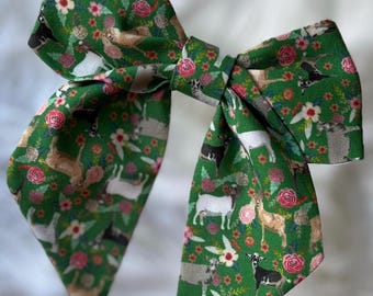 May include: A large fabric hair bow featuring a vibrant green background with a repeating pattern of various goats in shades of white, brown, and black, interspersed with small pink and white floral blossoms throughout the entire textile surface.