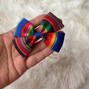 May include: A colorful bow with a striped pattern in shades of blue, red, green, and yellow. The bow is held in a hand and has a blue center knot. The bow is likely a hair accessory.