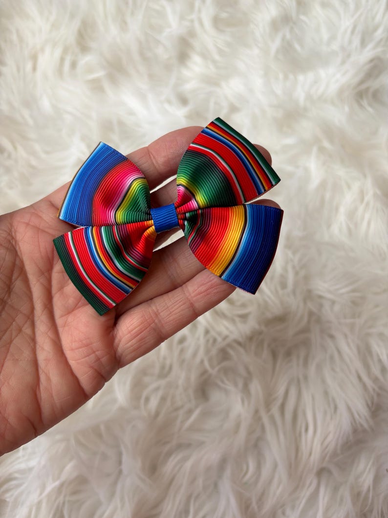 May include: A colorful bow with a striped pattern in shades of blue, red, green, and yellow. The bow is held in a hand and has a blue center knot. The bow is likely a hair accessory.