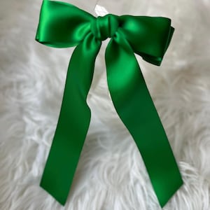 以下が含まれることがあります： 長く垂れ下がった鮮やかな緑色のサテンリボン。リボンは中央で結ばれており、クラシックな形をしています。鮮やかな緑色と滑らかな質感は、ギフト包装やヘアアクセサリーなど、装飾用のアイテムを示唆しています。