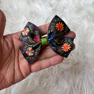 May include: A black hair bow with a colorful floral pattern. The bow features a green center and is held in a hand against a white, fluffy background. The floral design includes red, yellow, blue, and green accents.