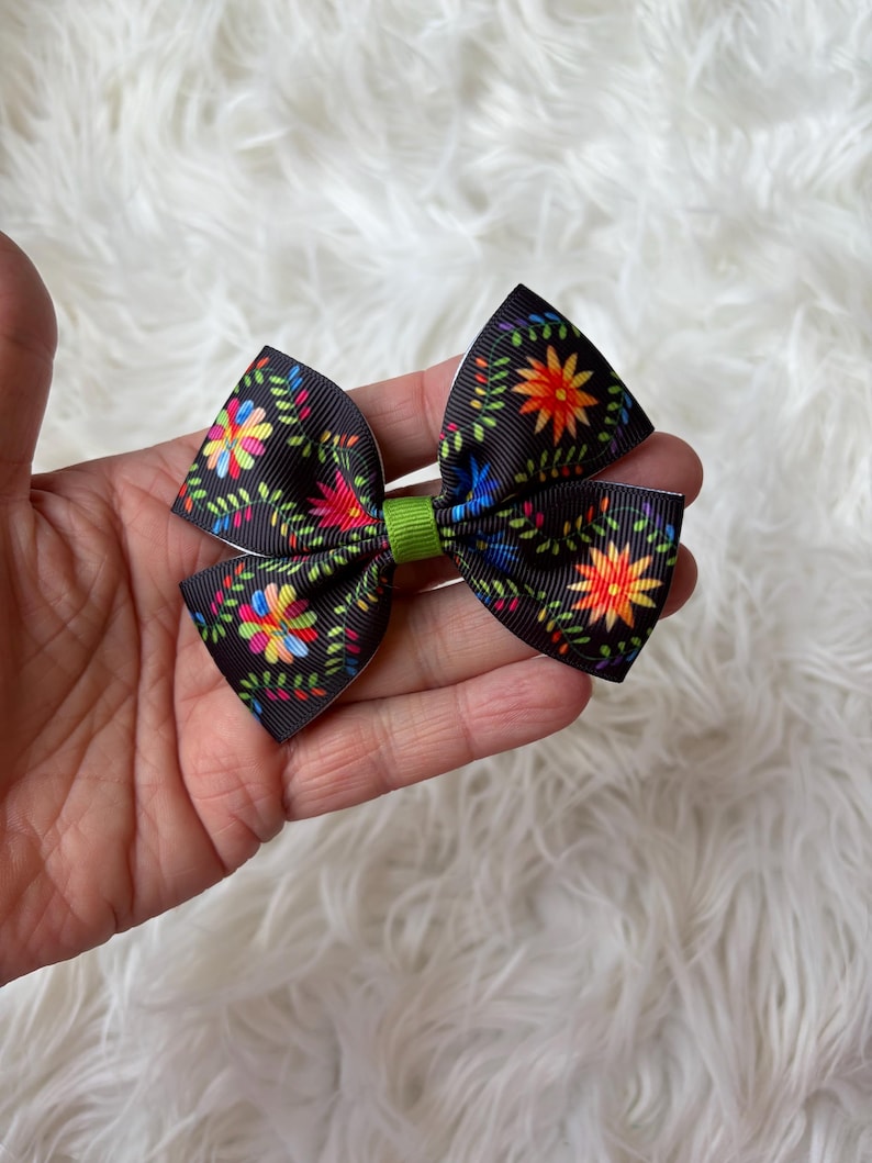 May include: A black hair bow with a colorful floral pattern. The bow features a green center and is held in a hand against a white, fluffy background. The floral design includes red, yellow, blue, and green accents.