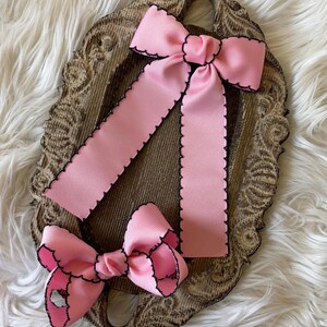 May include: Three pink hair bows with black trim are displayed on a decorative, ornate, brown tray. The bows are made of a grosgrain ribbon with a scalloped edge. The tray sits on a white, fluffy surface.