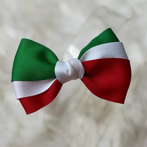 May include: A handmade bow featuring the colors of the Italian flag: green, white, and red. The bow is made of grosgrain ribbon and has a knot in the center. The bow is likely for hair accessories or decorative purposes.