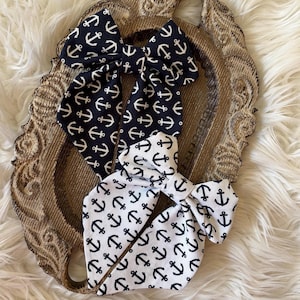 May include: Two fabric hair bows featuring a repeating anchor pattern. One bow is dark navy blue with white anchors, and the other is white with dark navy blue anchors. Both bows have long, pointed tails and a central knot detail made of fabric.