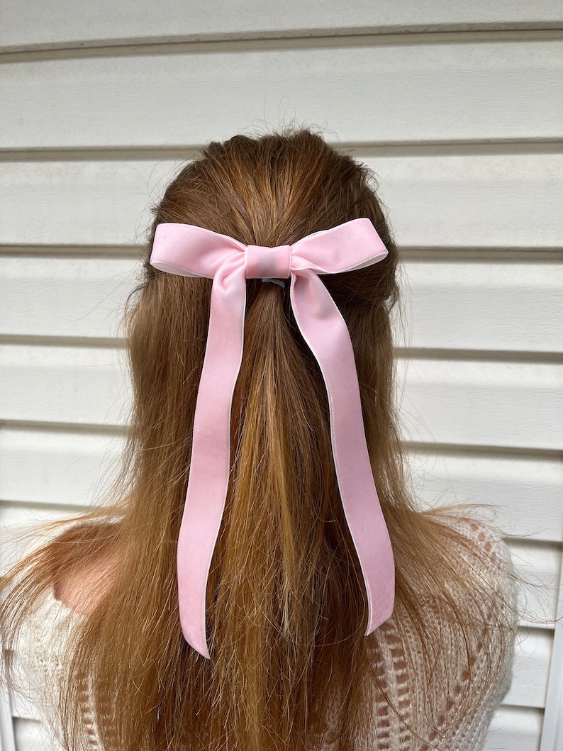May include: A pink velvet ribbon bow hair tie. The bow is tied in a simple knot and the ends of the ribbon are left long.