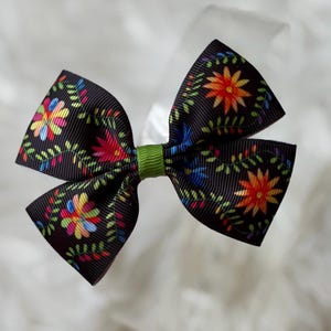 May include: A black hair bow with a floral pattern in red, orange, blue, and green. The bow is tied with a green ribbon in the center. The bow is approximately 4 inches wide.