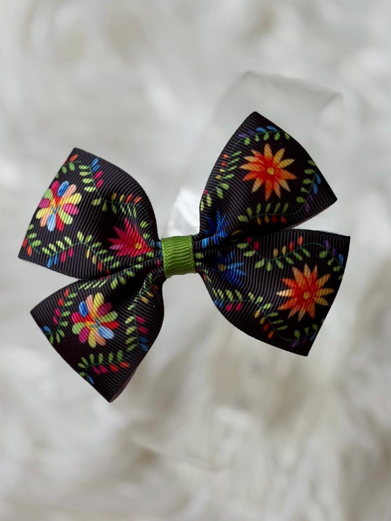 May include: A black hair bow with a floral pattern in red, orange, blue, and green. The bow is tied with a green ribbon in the center. The bow is approximately 4 inches wide.