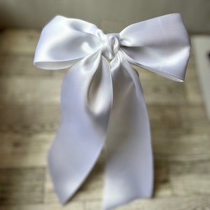 May include: A white satin ribbon tied into a large bow. The bow is tied in a way that the tails of the ribbon are long and flowing.