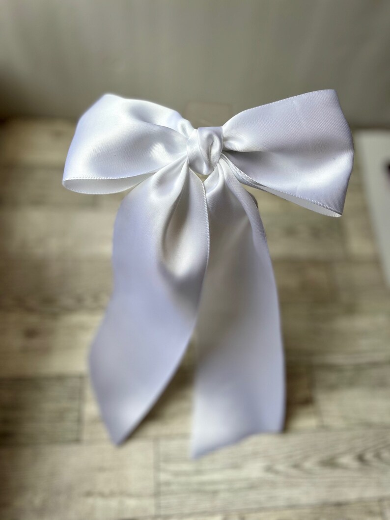 May include: A white satin ribbon tied into a large bow. The bow is tied in a way that the tails of the ribbon are long and flowing.