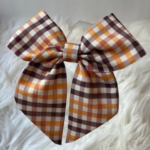 May include: A fabric hair bow featuring a multi-colored gingham pattern in shades of dark brown, light brown, orange, and white. The bow has two wide, pointed tails extending downward and a central knot detail holding the pleated fabric together.