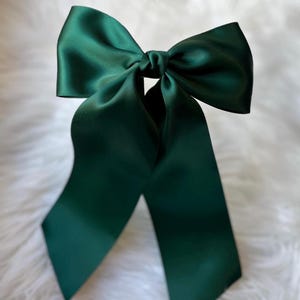 May include: A large, dark green satin bow with long, flowing tails. The bow is tied in the center, creating a full, elegant shape. The fabric has a slight sheen, and the bow is set against a soft, white background.