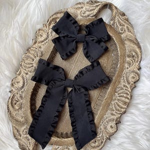 May include: Two black fabric hair bows with ruffled edges. The bows are displayed on a wooden tray with a decorative design.