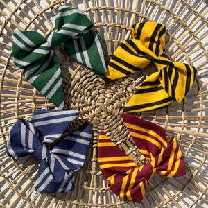 May include: Four fabric hair bows featuring striped patterns in distinct color combinations: green and silver, yellow and black, navy blue and silver, and maroon and gold. Each bow is constructed with multiple folded loops and pointed ends.