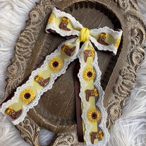 May include: A hair bow featuring a yellow ribbon base with embroidered brown cowboy boots and yellow sunflowers. The ribbon is edged with white scalloped lace trim and tied in the center with a solid yellow ribbon knot, layered over a brown ribbon.
