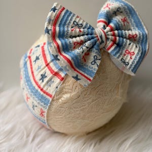 Puede incluir: Una diadema para bebé color crema con un lazo grande. El lazo presenta un diseño patriótico con rayas rojas y azules, estrellas y patrones de lazo sobre un fondo blanco. La diadema es de encaje.