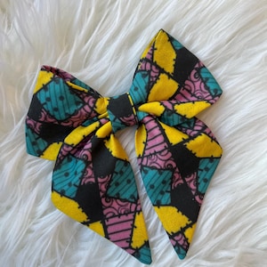 Op de afbeelding: Een grote stoffen strik met een patchwork ontwerp in geel, turquoise, zwart en roze. De strik is in het midden geknoopt, met de stof symmetrisch gerangschikt. Het ligt op een pluizige witte achtergrond.