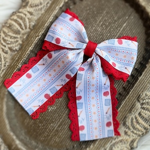 May include: A hair bow featuring a white ribbon with a blue gingham pattern, red apples, pencils, and the letters ABC. The bow is layered with a solid red scalloped lace trim and secured in the center with a solid red ribbon knot.
