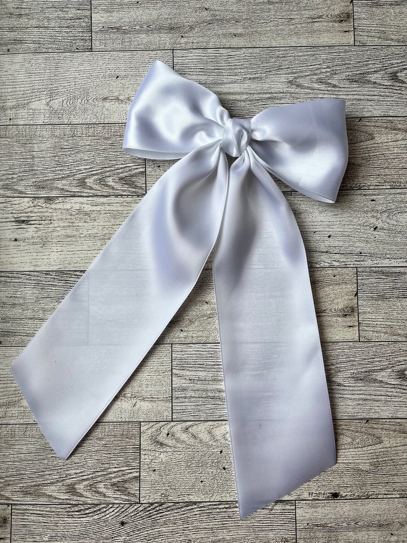 May include: A large white satin bow with long tails. The bow is tied in a knot and the tails are hanging down.