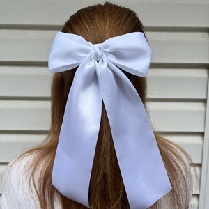 May include: A white satin ribbon bow hair tie. The bow is tied in a large, loose knot and the tails of the ribbon are long and flowing.