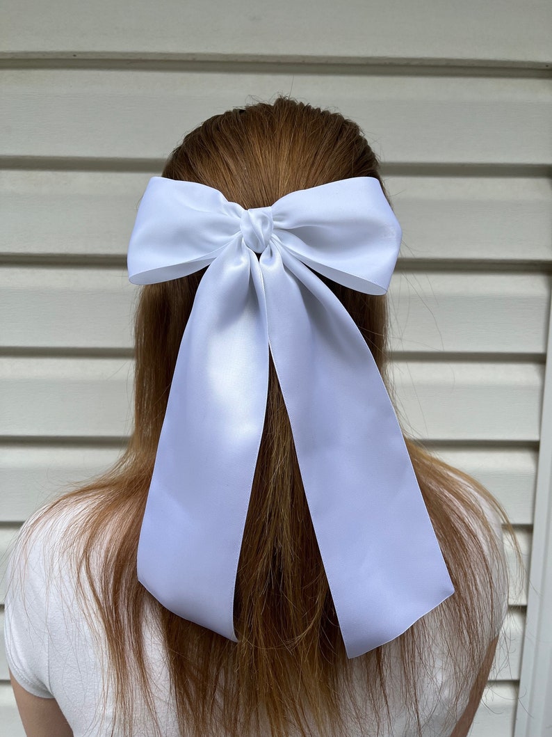 May include: A white satin ribbon hair bow. The bow is tied in a large, loose knot and the tails of the ribbon are long and flowing.