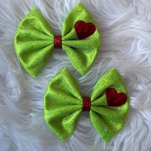Peut inclure: Deux nœuds pour cheveux vert lime avec des centres rouges pailletés et des accents de cœur rouges. Les nœuds sont faits d'un tissu scintillant et sont posés sur un fond blanc et moelleux. Parfaits pour coiffer.