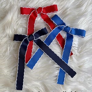 Pode incluir: Laços de fita vermelhos, azul marinho e azul royal com bordas onduladas brancas são exibidos em uma superfície branca e fofa. Os laços são amarrados no centro. O texto na parte inferior diz "More Colors Available!"