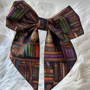 Pode incluir: Uma gravata borboleta grande e decorativa com uma impressão detalhada de uma estante de biblioteca. O tecido apresenta uma variedade de lombadas de livros em tons de vermelho, verde, roxo e dourado, com um fundo escuro. A gravata borboleta inclui um design de relógio.