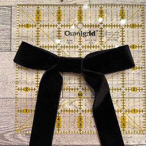 May include: A black velvet bow tie on a clear ruler with yellow markings. The ruler is marked in inches and has a 60 degree angle.