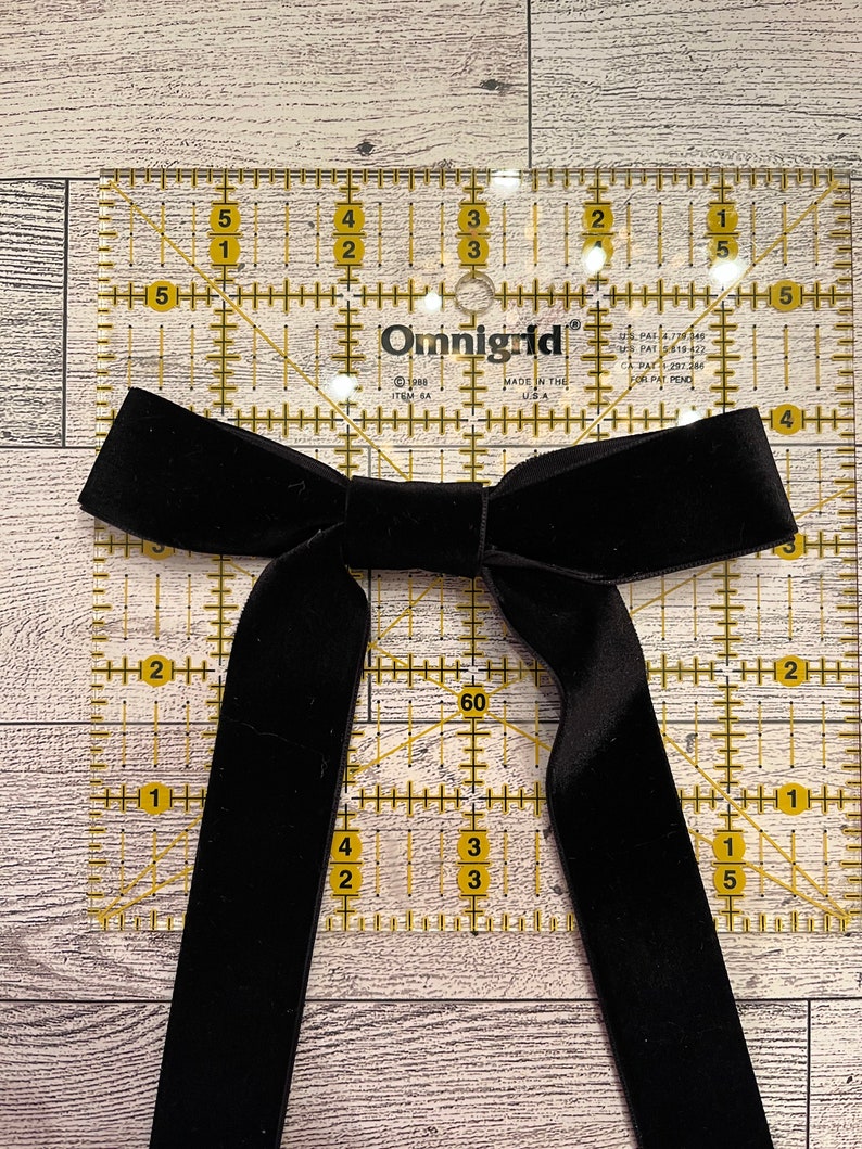 May include: A black velvet bow tie on a clear ruler with yellow markings. The ruler is marked in inches and has a 60 degree angle.
