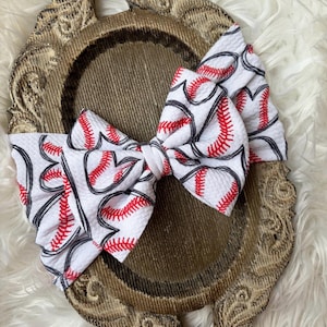 May include: A large, fabric hair bow featuring a baseball design. The bow is white with a repeating pattern of baseballs, each with red stitching and black outlines. The bow is tied in the center and sits on a decorative, ornate, gold-toned tray.