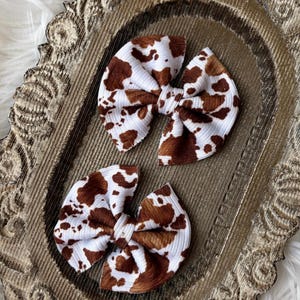 May include: Two hair bows made of a white ribbed fabric featuring a brown cow print pattern. Each bow is constructed with a central knot detail and gathered fabric loops, displaying a consistent texture and color scheme across both items.