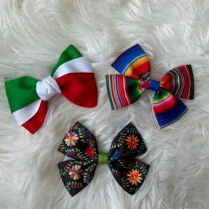 May include: Three decorative hair bows. One bow features the colors of the Italian flag. Another bow has a colorful striped pattern. The third bow is black with a floral design and a green center.