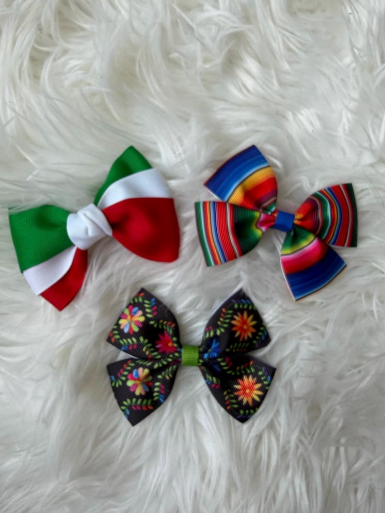 May include: Three decorative hair bows. One bow features the colors of the Italian flag. Another bow has a colorful striped pattern. The third bow is black with a floral design and a green center.