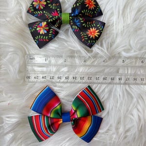 May include: Two decorative hair bows. The top bow is black with colorful floral patterns and a green center. The bottom bow features a vibrant striped design in red, blue, green, and yellow. A ruler is placed next to the bows.