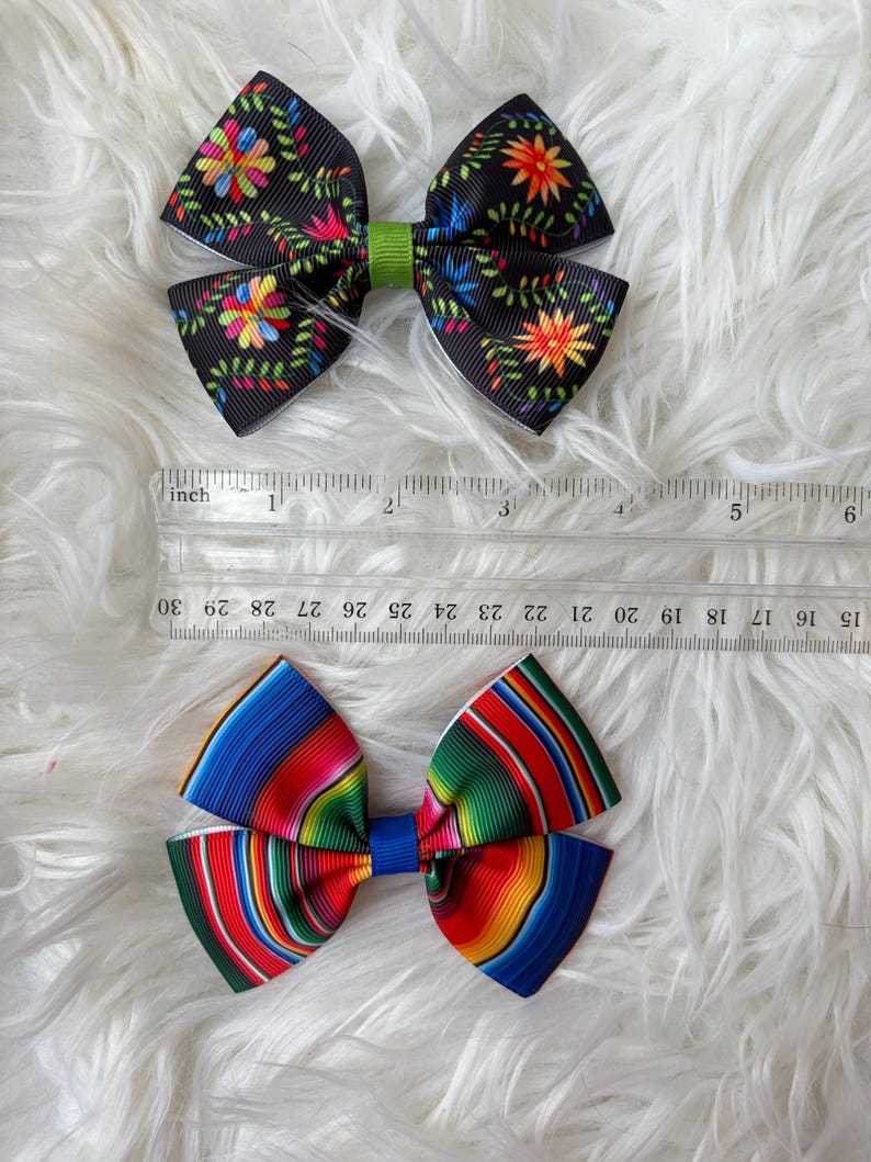May include: Two decorative hair bows. The top bow is black with colorful floral patterns and a green center. The bottom bow features a vibrant striped design in red, blue, green, and yellow. A ruler is placed next to the bows.