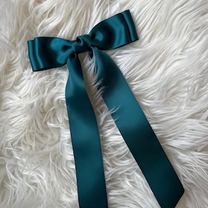May include: A teal satin hair bow with long, flowing tails. The bow is tied in the center and rests on a fluffy, white faux fur surface. The bow's color is a deep, rich teal.
