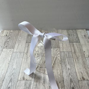 Może przedstawiać: Przezroczyste plastikowe pudełko z białą satynową wstążką zawiązaną w kokardę. Pudełko stoi na jasnym drewnianym podłożu.