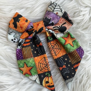 May include: A quilted fabric hair bow featuring a patchwork pattern of Halloween motifs including black cats, orange pumpkins, white ghosts, spiders, spiderwebs, and stars in shades of orange, black, green, and purple with a stitched texture.